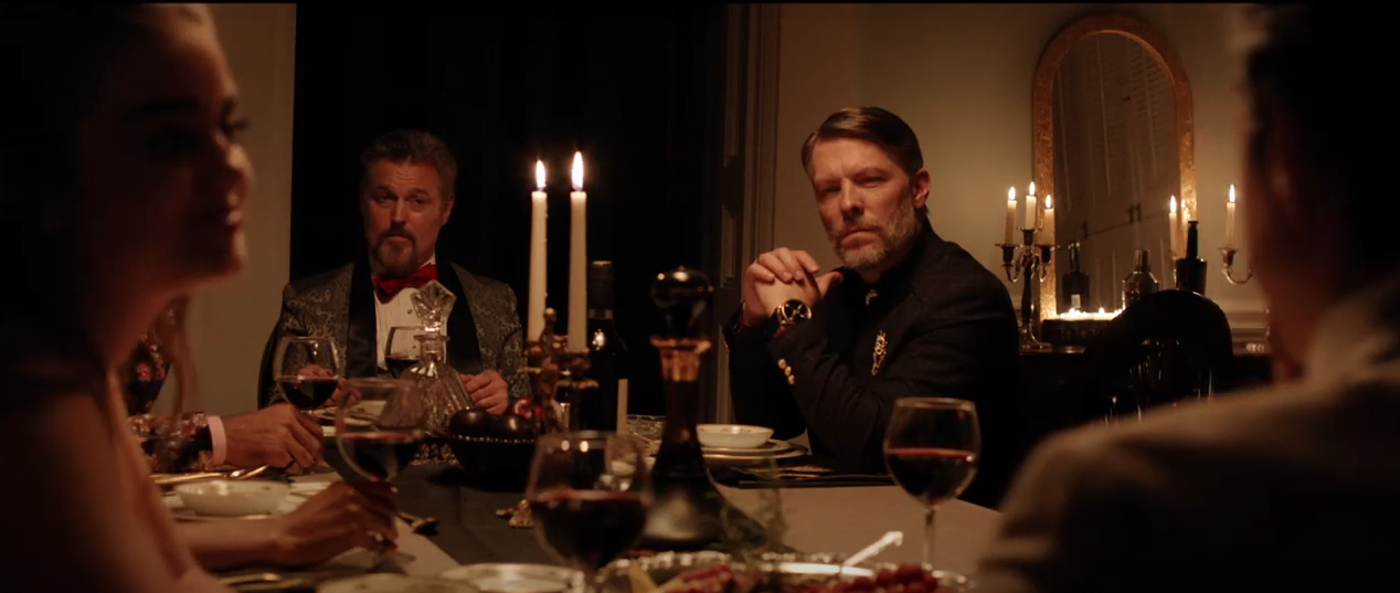 From the film 'The Dinner Party': Two men stare inquisitively and ominously, across a fine dining table, from a woman on the left of the screen, looking at her husband on the right.