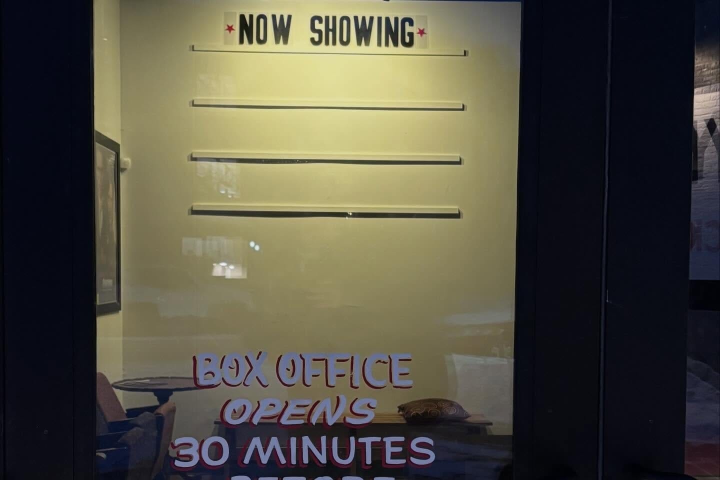 Trylon Cinema's ticket booth, with a "Now Showing" sign, above empty movie listings, behind a glass decal that says "Box Office Opens 30 Minutes."